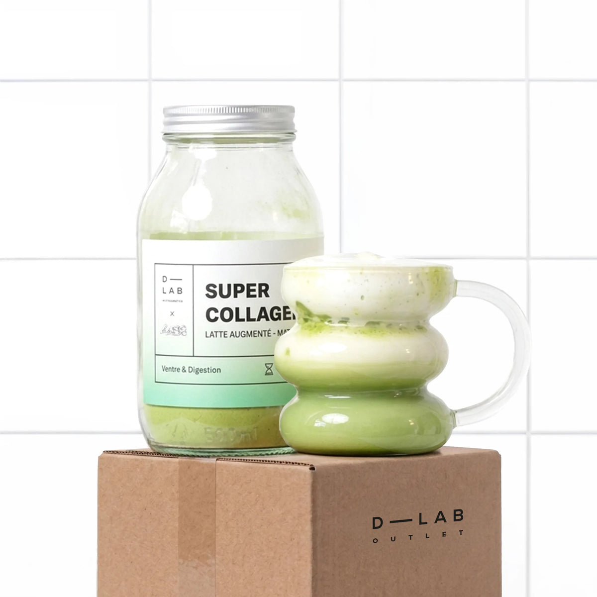 Matcha Latte au Collagène D-LAB, pot en verre et tasse de latte matcha, sur le carton D-LAB Outlet, devant un mur carrelé blanc